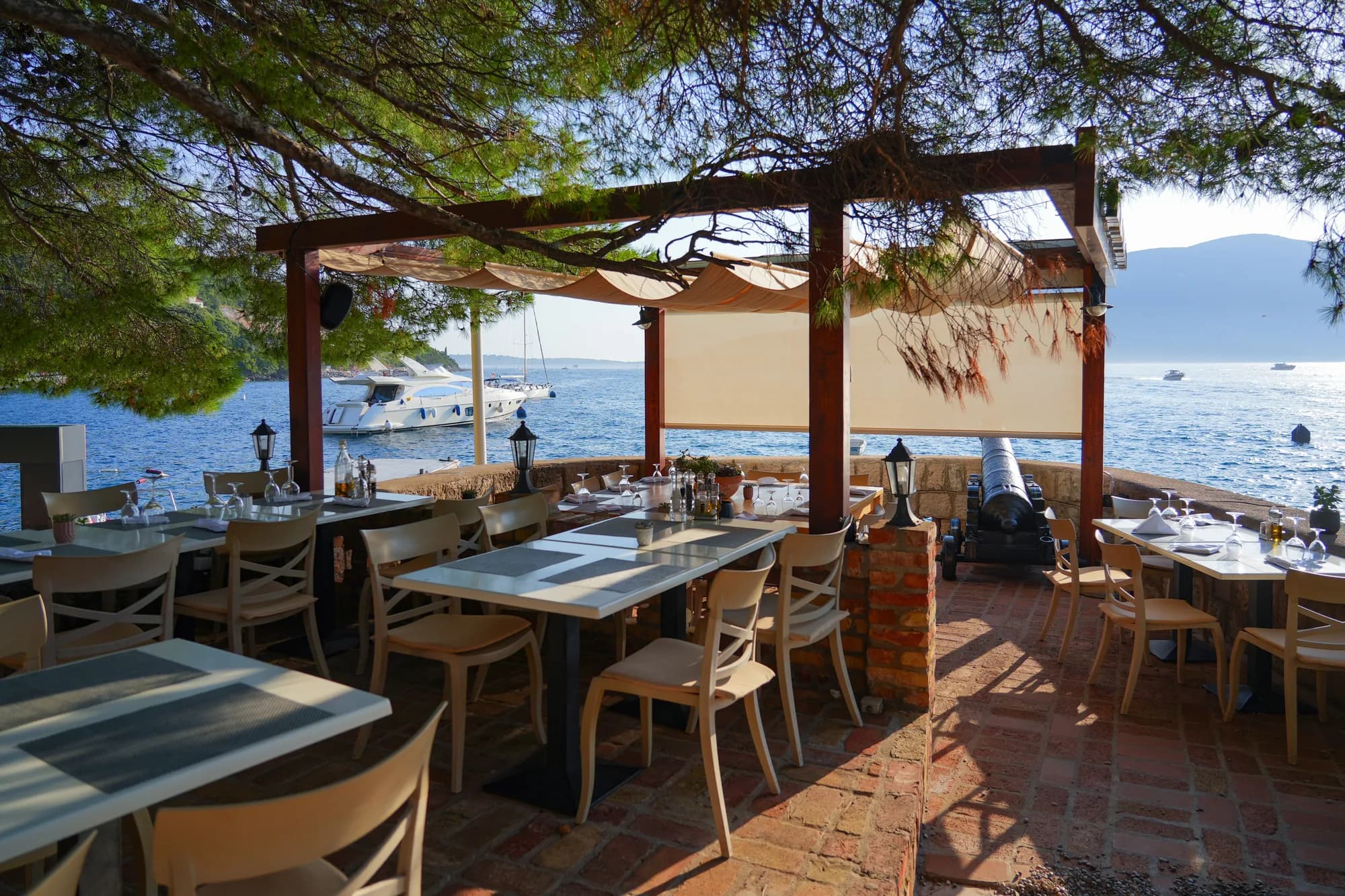 A restaurant terrace at the water in Rose, tables laid under pines and an awning, boats at anchor beyond — the reason most people make the crossing