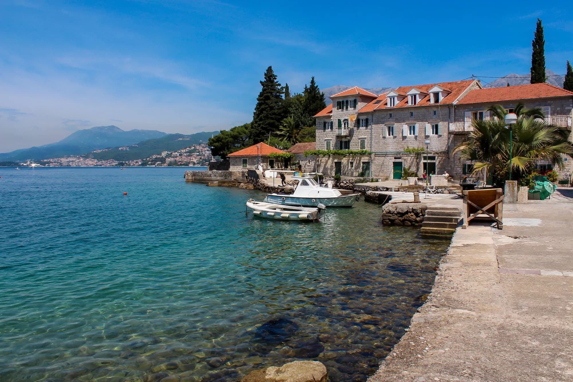 The waterfront at Rose: stone houses with red roofs at the edge of the water, small boats moored off the quay and clear shallows over pebbles, the shore of Herceg Novi across the mouth of the bay