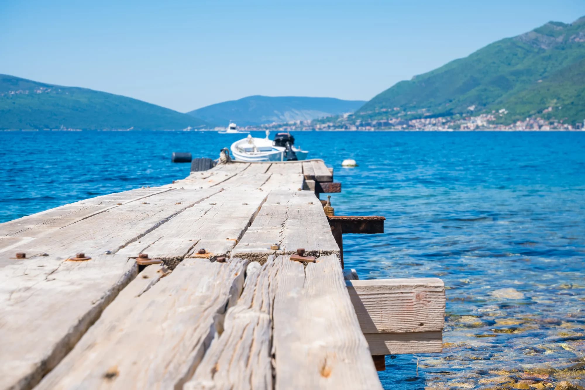 A weathered wooden jetty running out over clear water at Rose, a small boat at its end and the far shore of the bay mouth behind