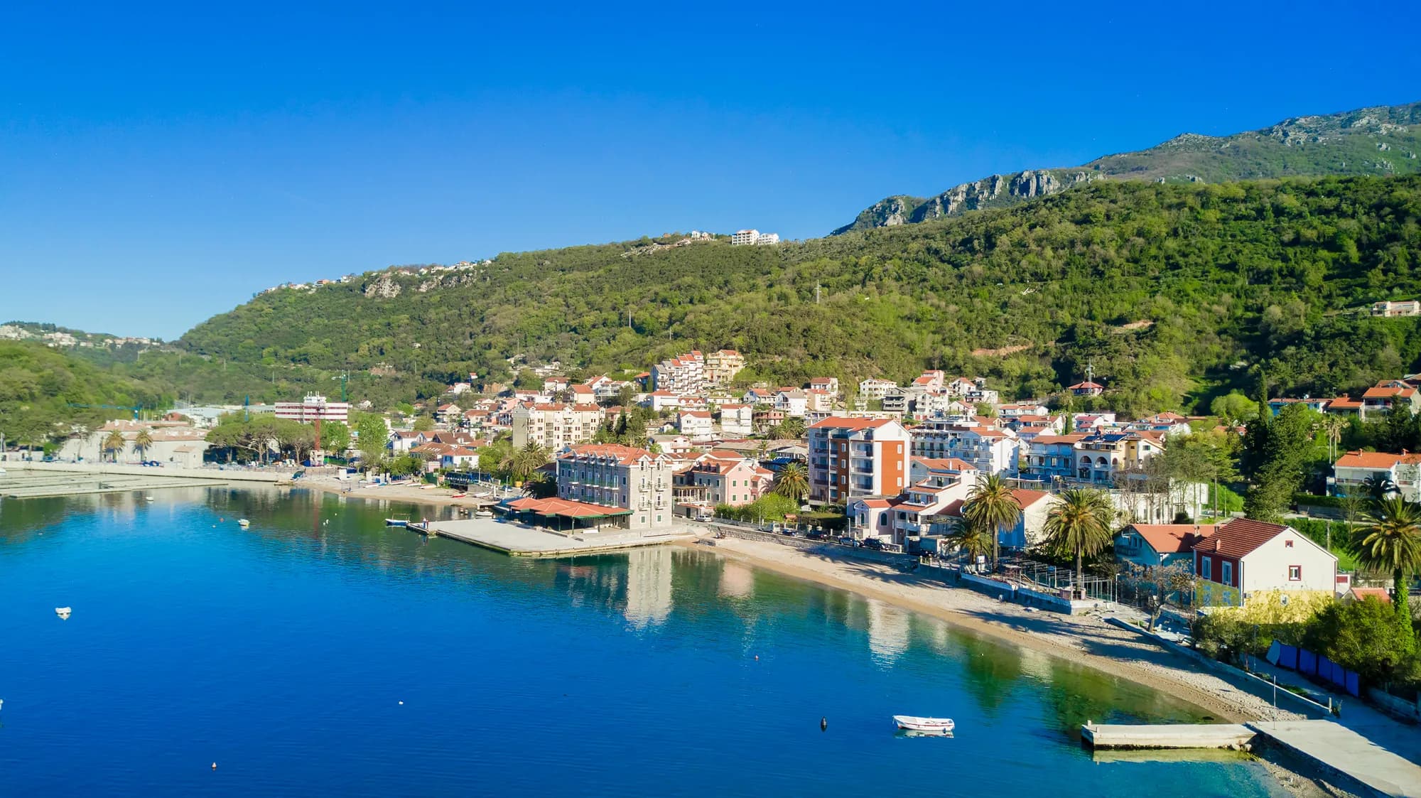 The shore between Herceg Novi and Meljine from the air: the strip of beach and the promenade along the water, the pier, and the wooded slope of the monastery hill behind the roofs