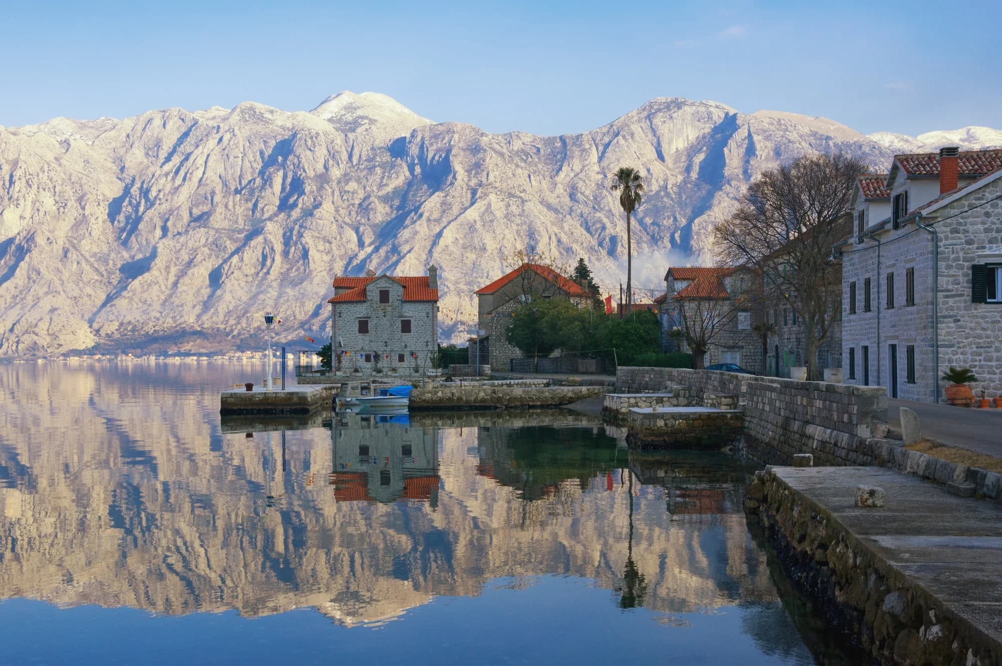 A stone house with a roof turret on a point at Donji Stoliv, a small boat in its stone basin, concrete platforms along the water and snow on the mountains across the bay
