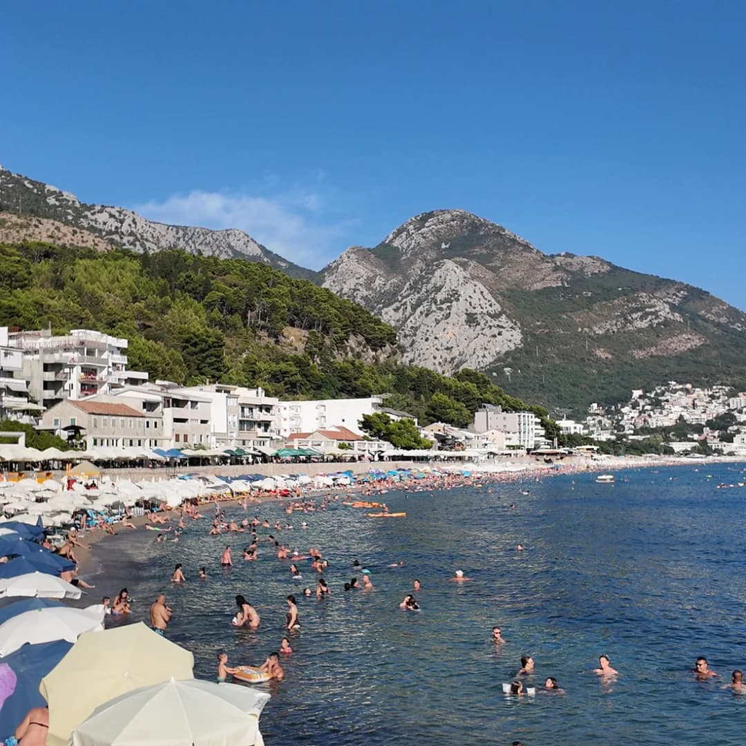 A wide sandy beach curving along the water with steep mountains rising directly behind it.