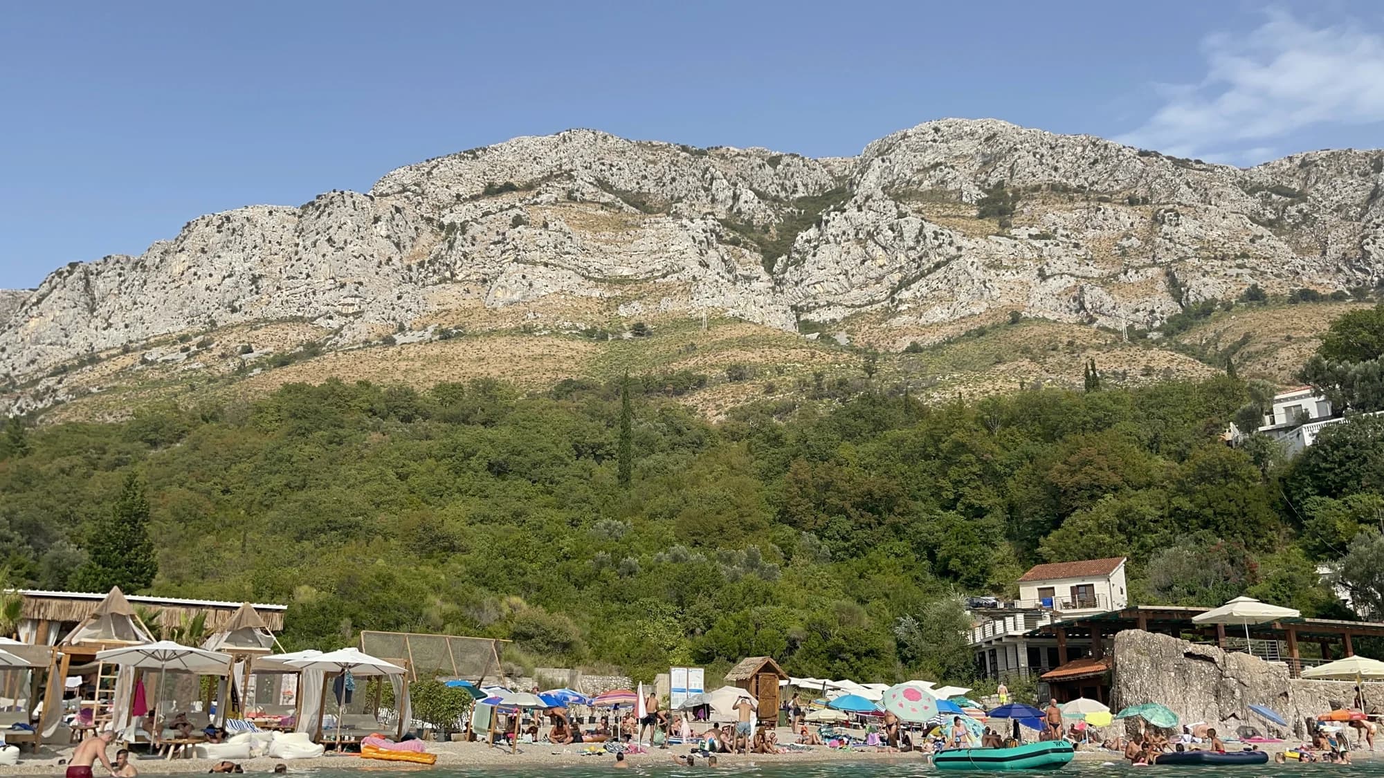 The line of cabanas and loungers along Drobni Pijesak, the bare limestone wall of the Paštrovići ridge rising directly behind