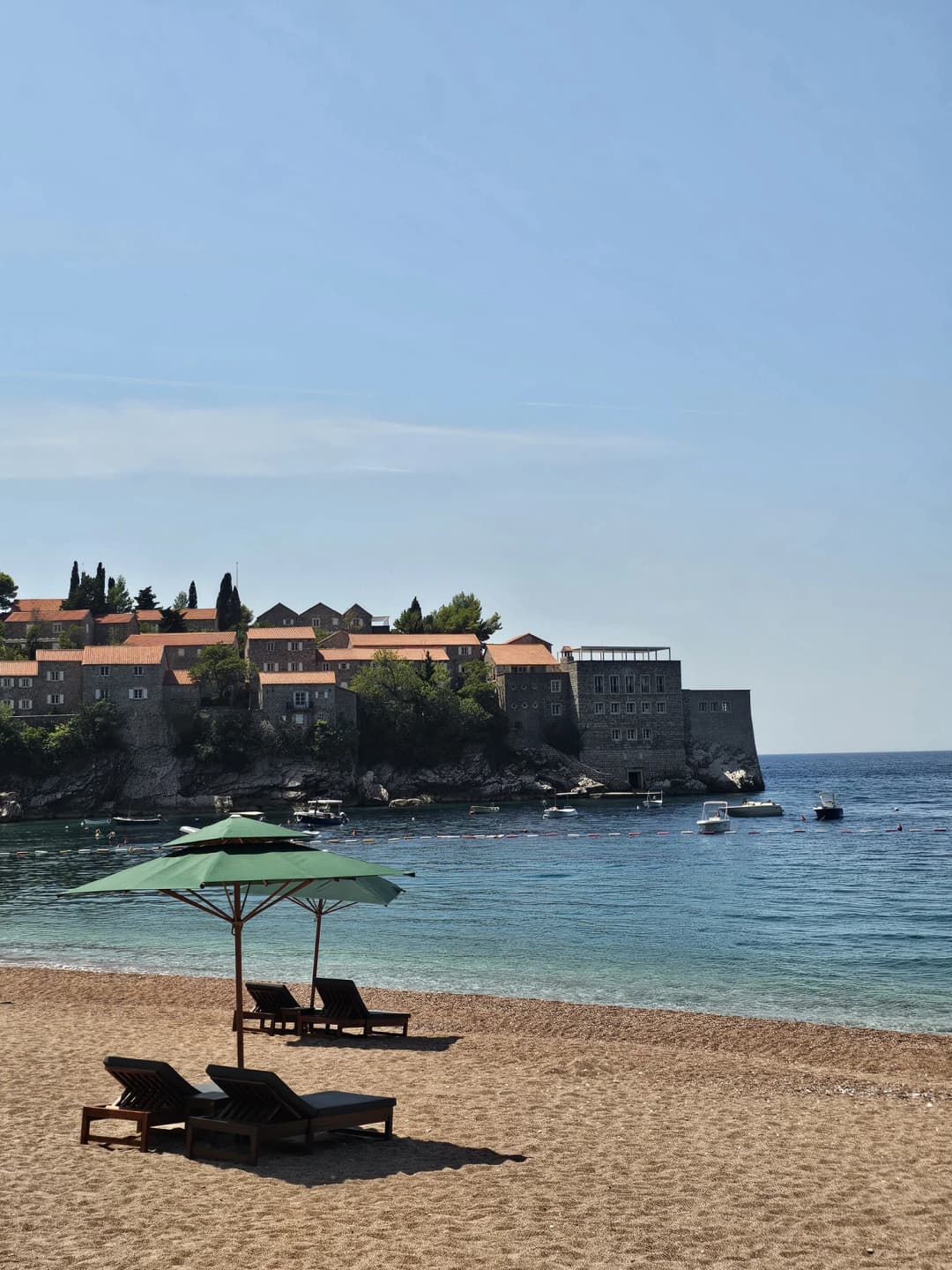 Sveti Stefan island above the pink pebble shore and green umbrellas of the public northern beach