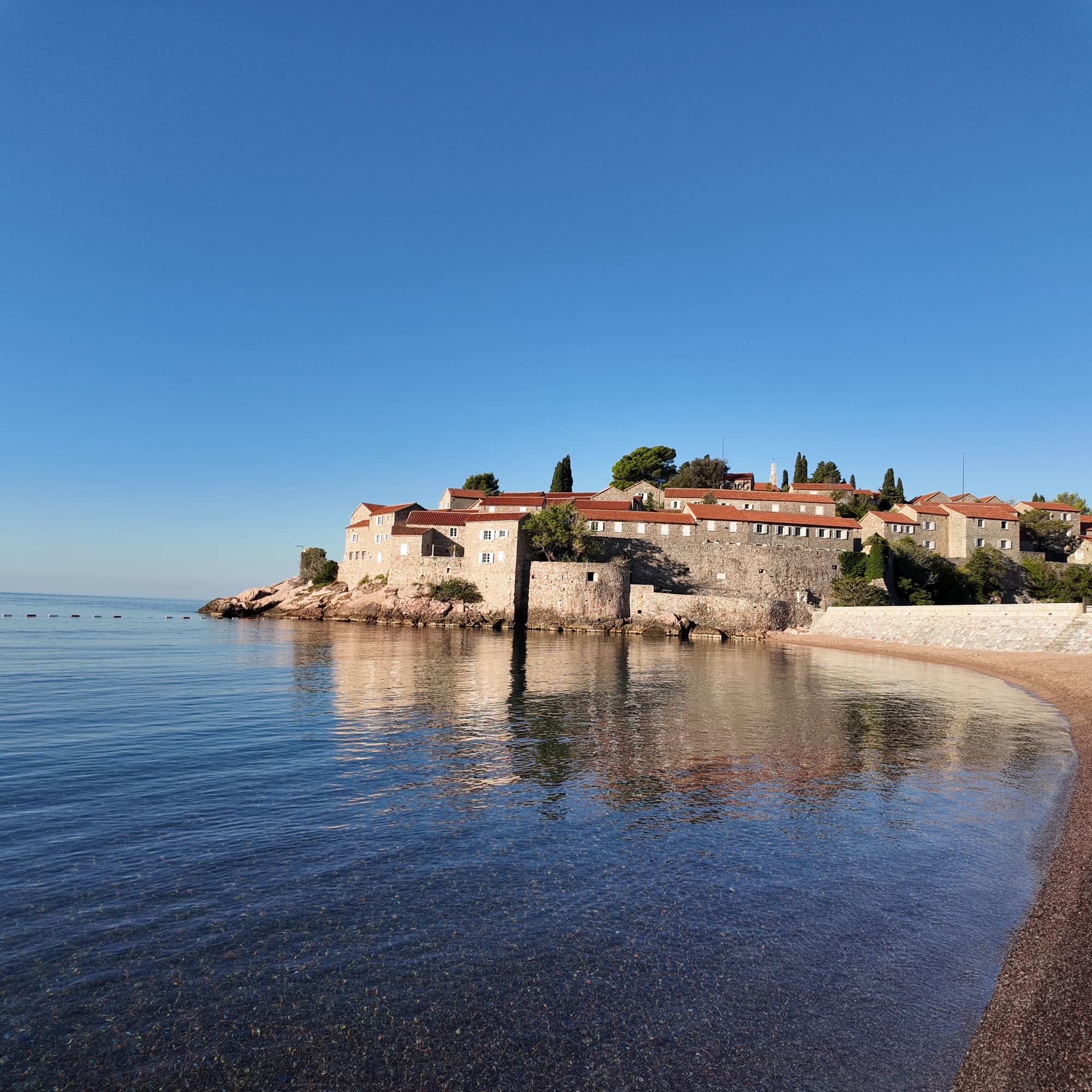 Sveti Stefan reflected in clear water beside the curving pebble beach in warm morning sun