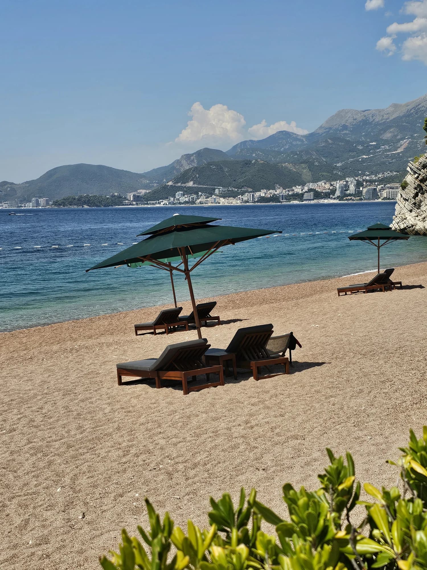 Green umbrellas and loungers on Sveti Stefan’s northern beach, looking across the turquoise water toward Budva