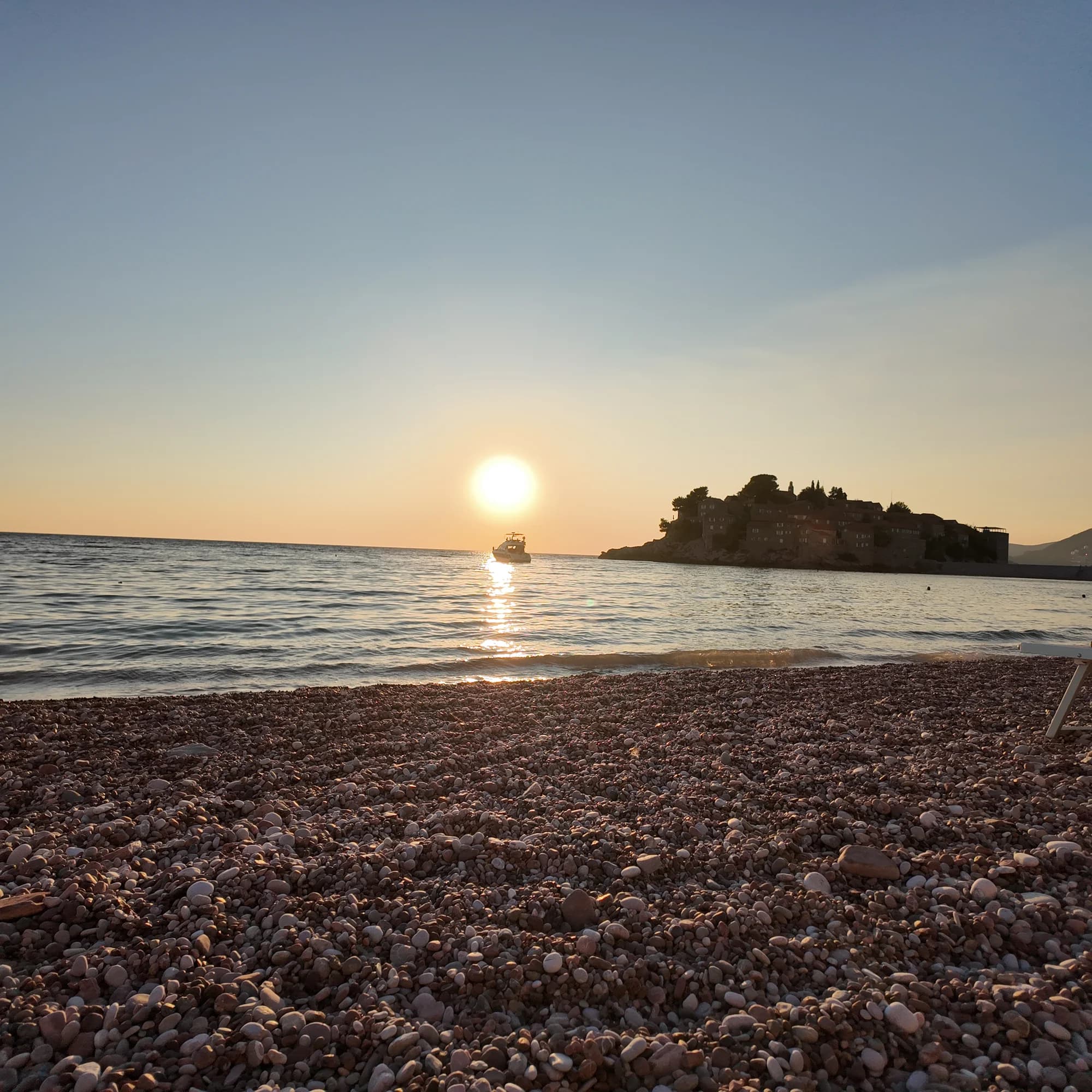 A boat crossing the sunlit water beside Sveti Stefan, with the northern beach’s pink pebbles in the foreground