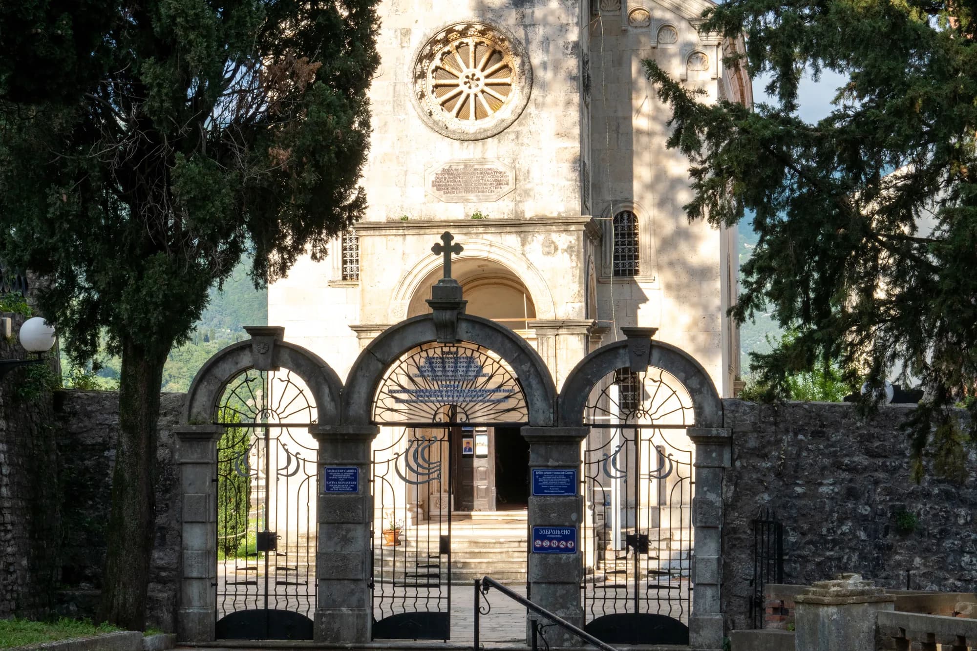 A church front with a rose window and a bell tower behind it, seen through the ironwork gates of its walled courtyard, cypresses on either side