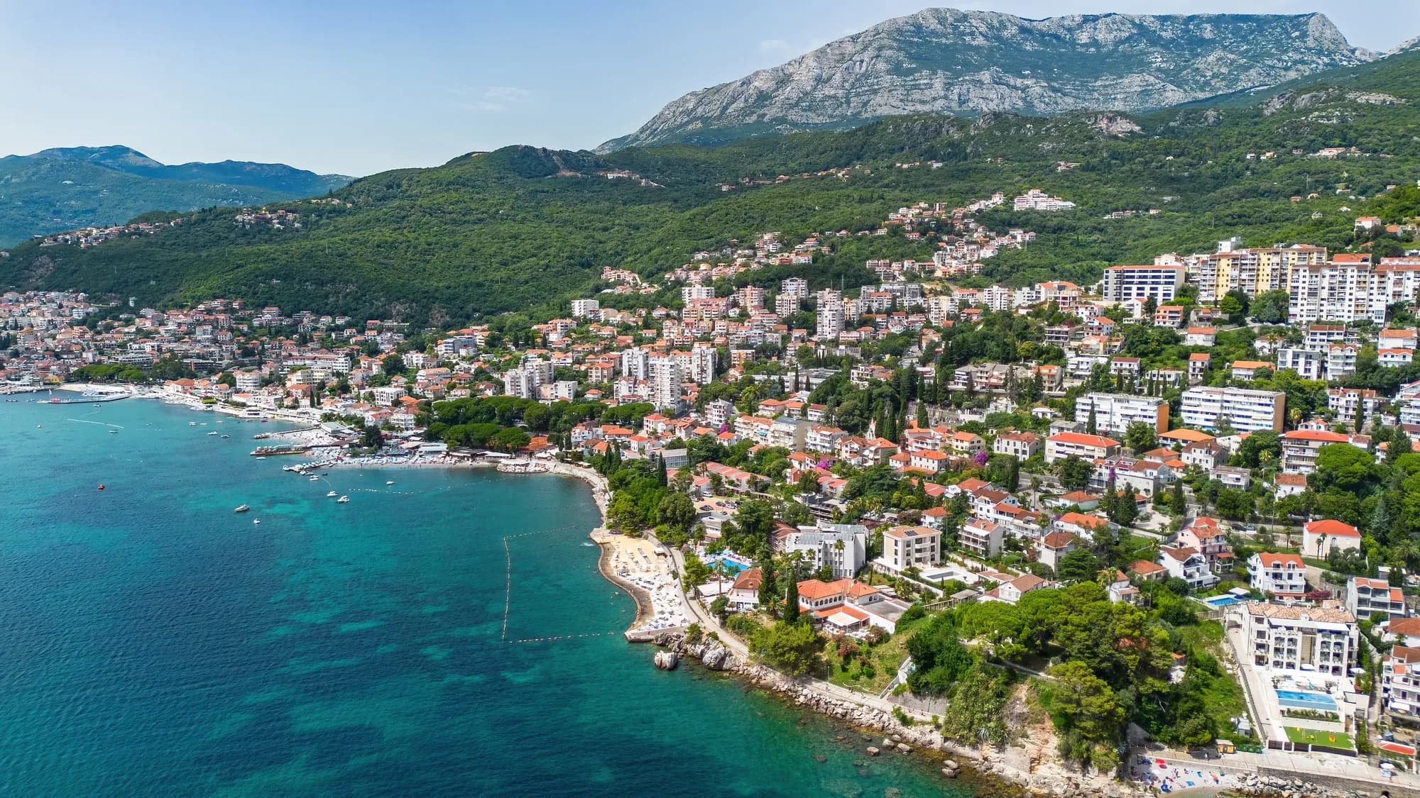 The Topla shore from the air: the pebble beach and swimming platforms at the water, apartment blocks climbing the slope behind, and the ridge above the town
