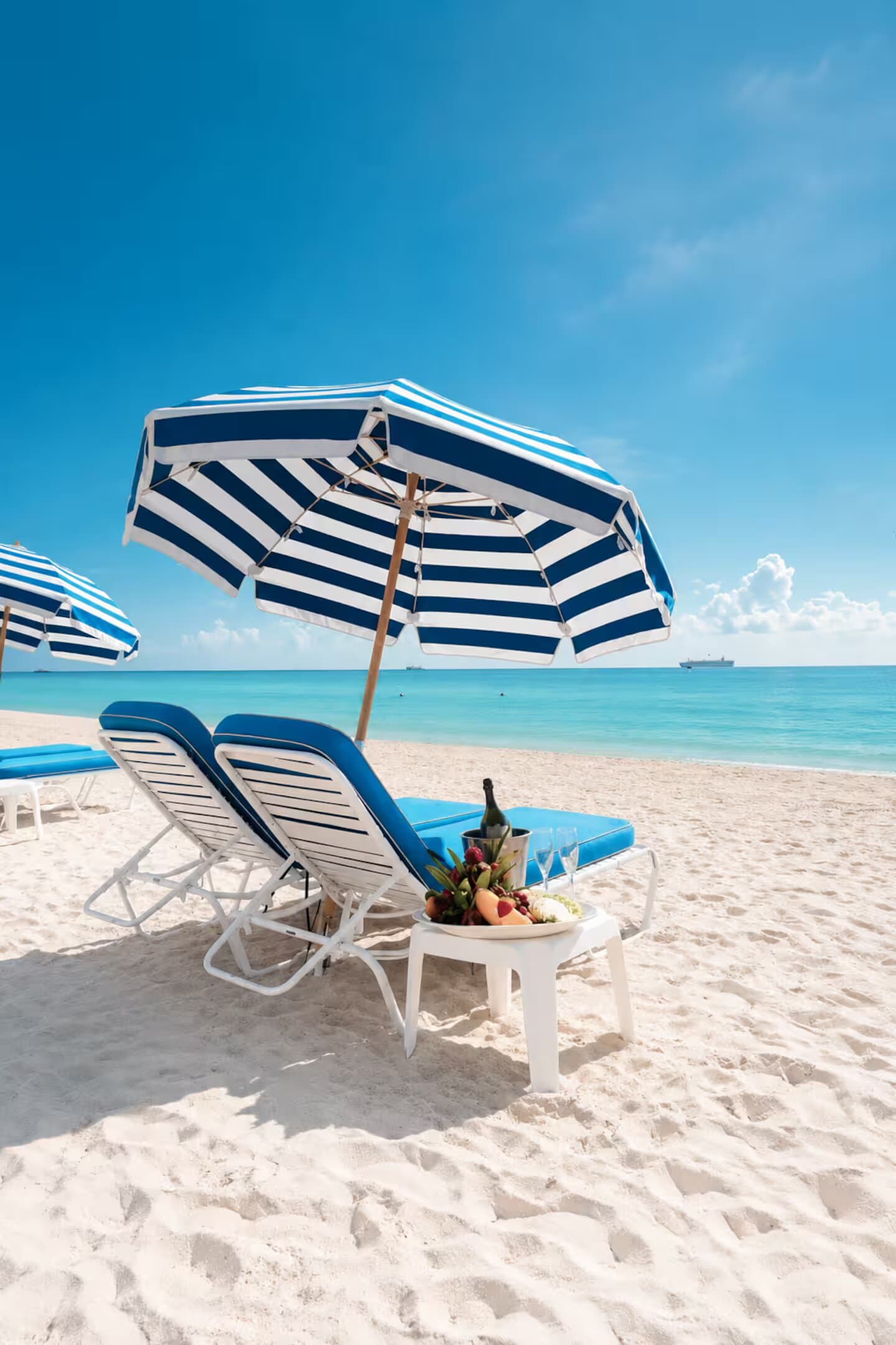 Two beach chairs and a striped umbrella set on the sand at the Bentley Beach Club, with champagne and fruit