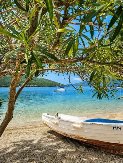 A wooden boat on the pebble shore under the oleander, sailboats in the bay