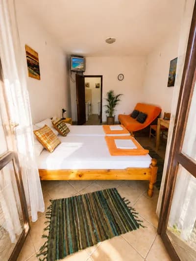 Bright studio with twin beds, orange sofa and tiled floor, doors open to the balcony