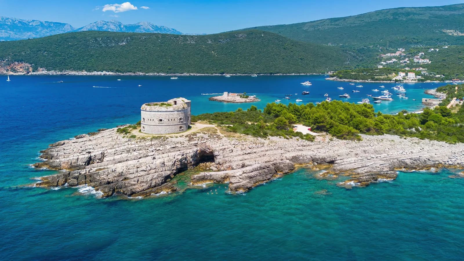 Aerial view of Fort Arza: a round grey stone fort with gun embrasures around its wall, standing on the tip of a bare limestone cape that shelves into clear turquoise water, with the wooded far shore of the strait and anchored boats beyond.