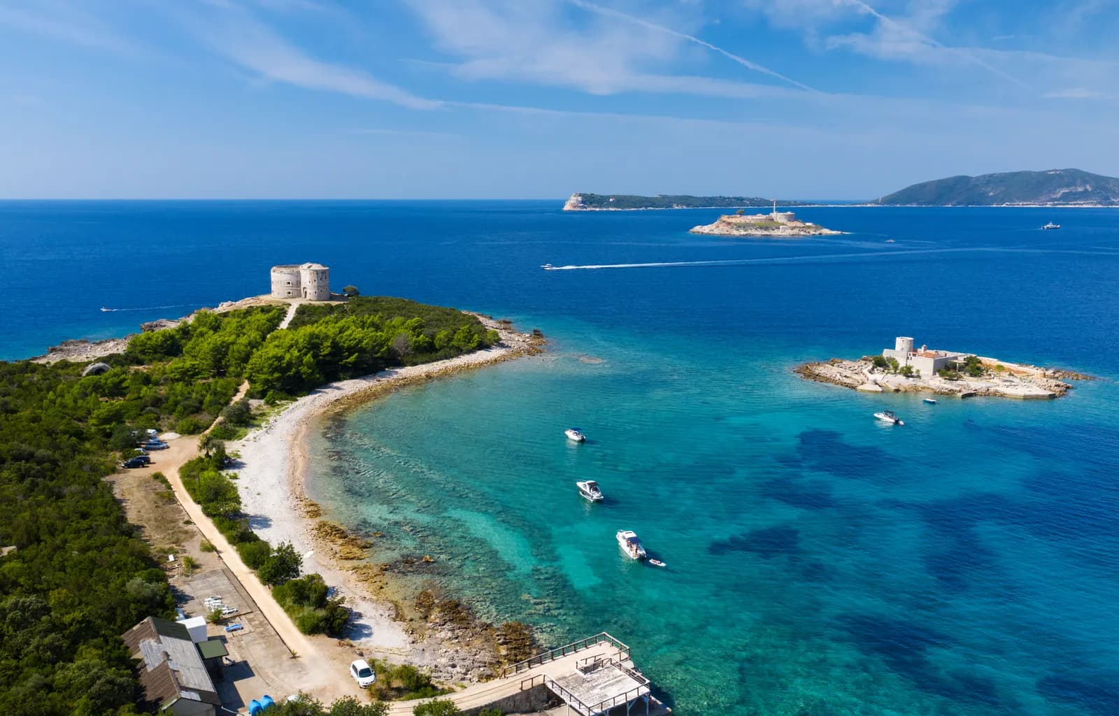 Aerial view over Cape Arza: the round Arza fort on its wooded headland at left, the fortified island of Mamula with its lighthouse in the middle distance, open sea and the far shore beyond, and small boats anchored in the turquoise bay between them.
