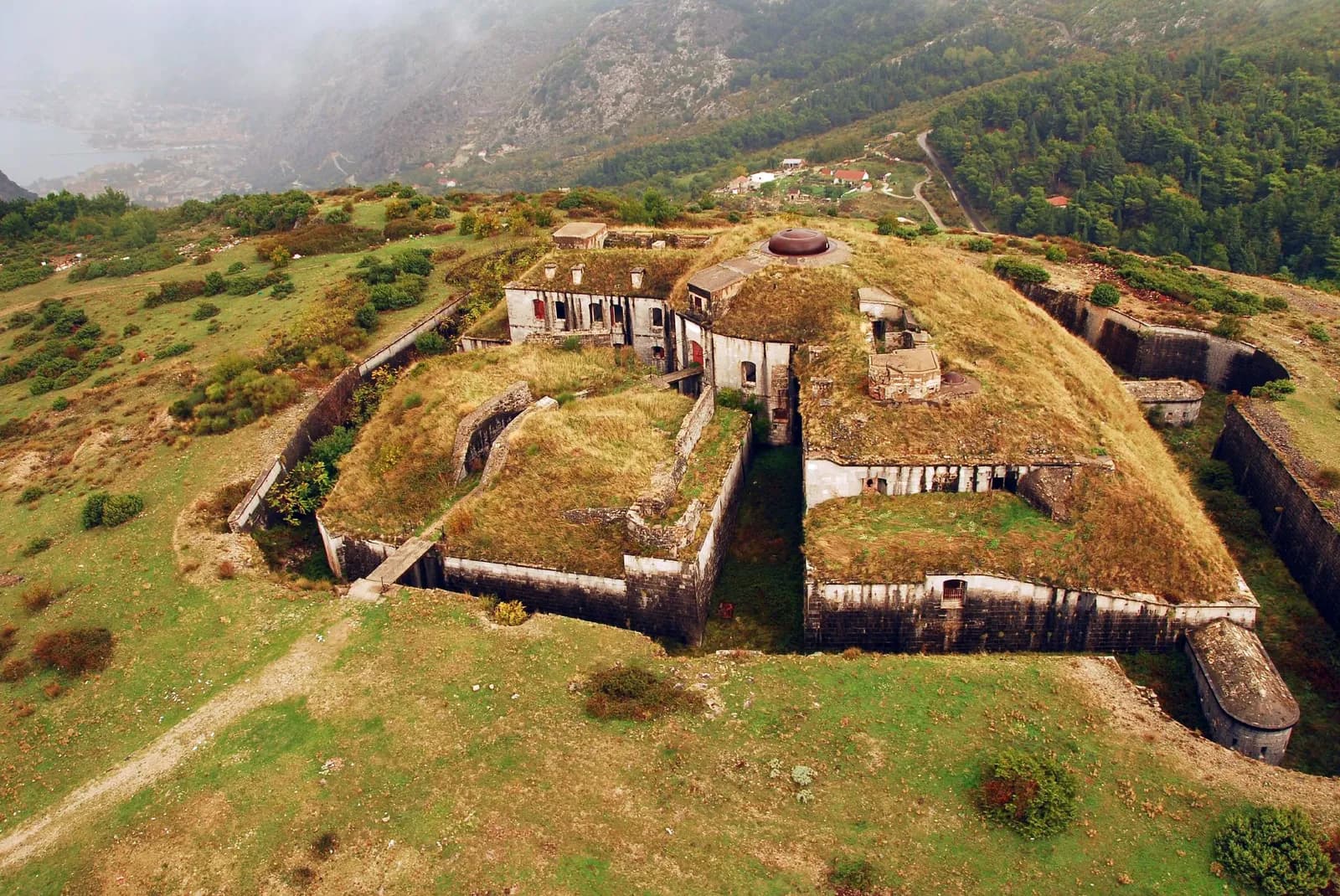 Aerial view of Fort Gorazda: a low polygonal fort set into a grassy hilltop, its roof covered in earth and dry grass with a rust-red armoured turret dome near the centre, a stone barrack range with rows of windows, dry ditches with angled walls around the outside, and the misty Bay of Kotor far behind.