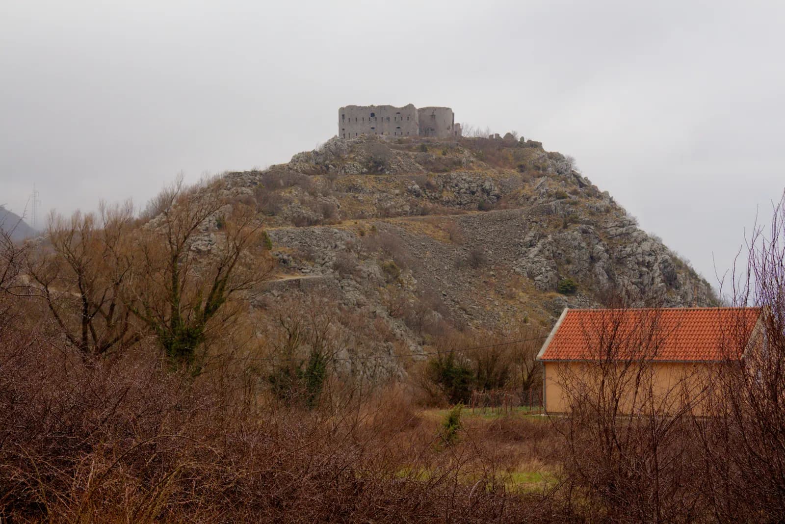 Fort Kosmač seen from the Budva–Cetinje road: a large rectangular grey stone barrack block standing roofless and empty on a bare hilltop, its two ranges of window openings showing sky through them, with scrub and mountain slopes around it.