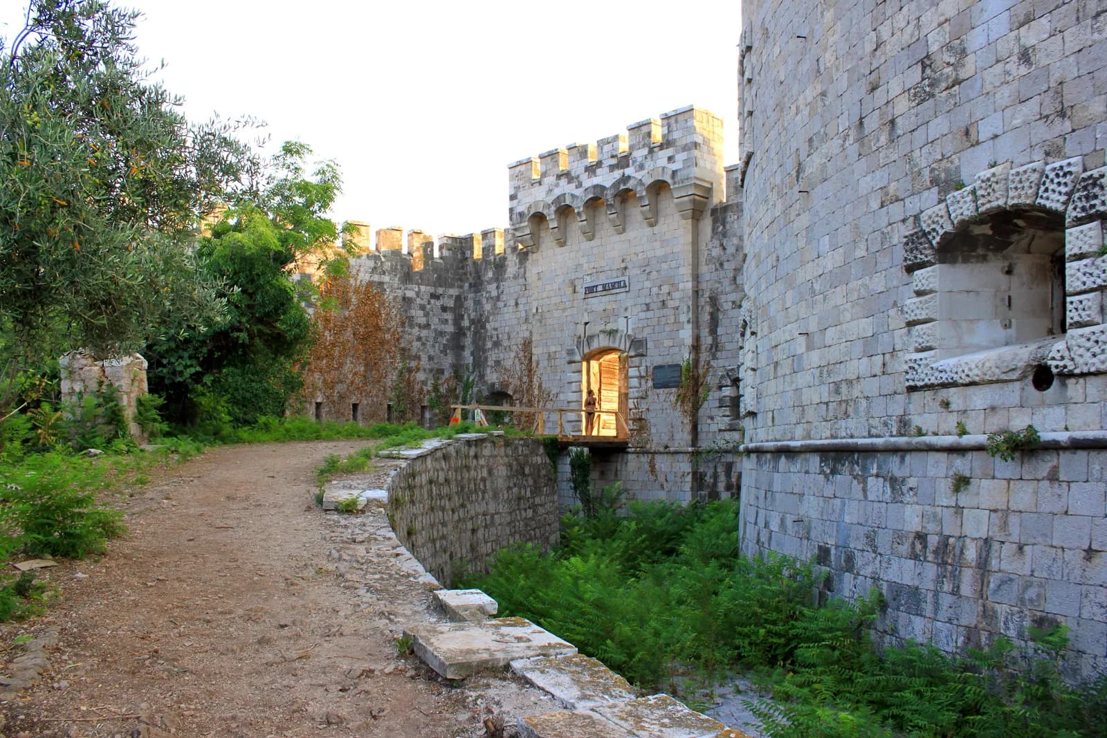 The entrance to Fort Mamula: a crenellated stone gatehouse with a timber bridge crossing a dry moat to its arched doorway, the massive curved ashlar wall of the circular fort rising at the right, and ferns growing in the moat below.