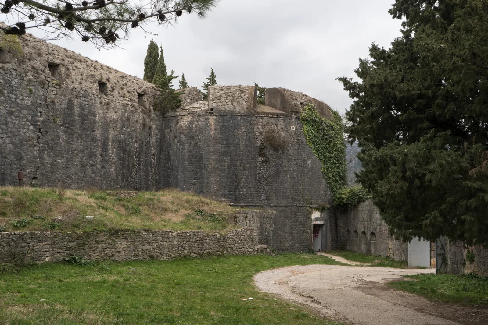 The curtain wall of Španjola fortress in 2024: a long, steeply battered grey stone wall with small rectangular gun embrasures, meeting a massive rounded bastion draped in ivy, with a track and grass in the foreground under an overcast sky.