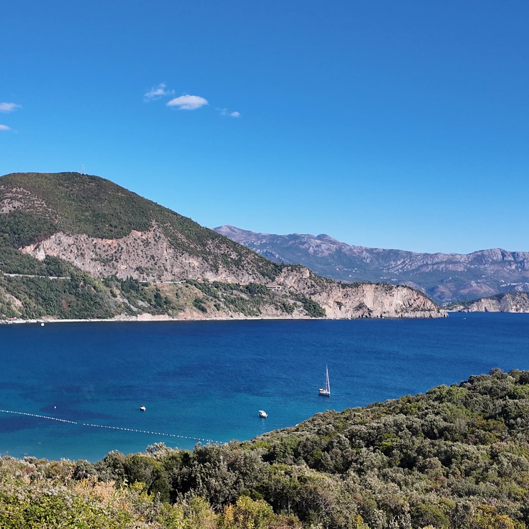 Jaz bay from the hill on the Budva side: the buoy line, a yacht at anchor in the blue water and the wooded headland with the highway cut closing the bay