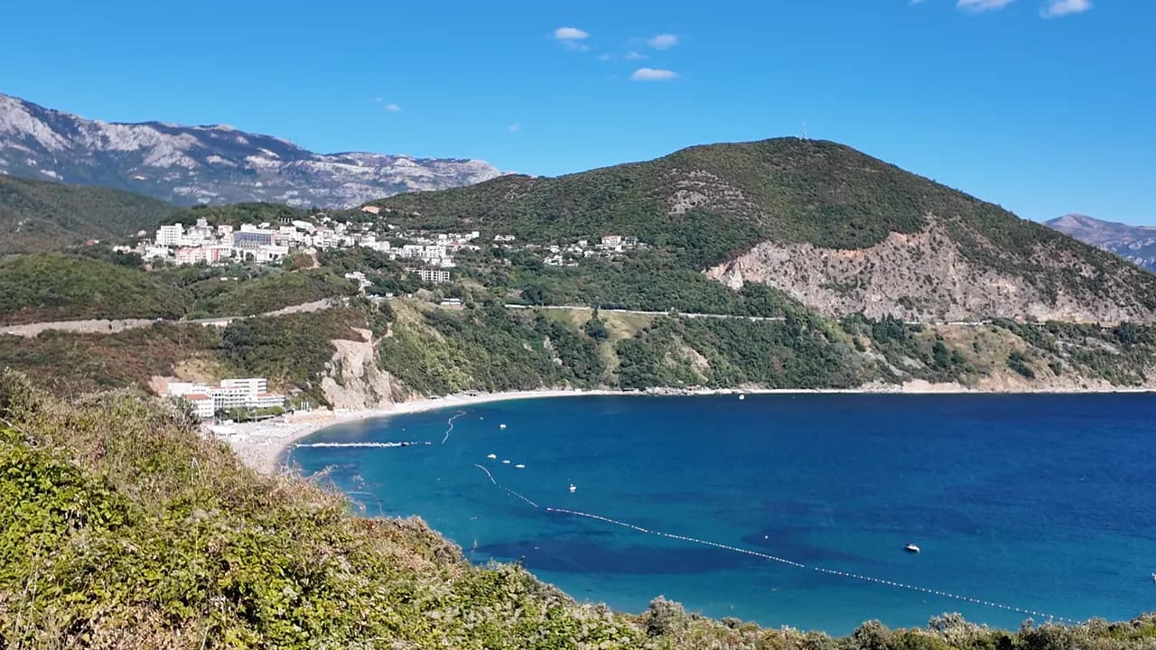 Jaz bay from the hill on the Budva side: the buoy line, a yacht at anchor in the blue water and the wooded headland with the highway cut closing the bay