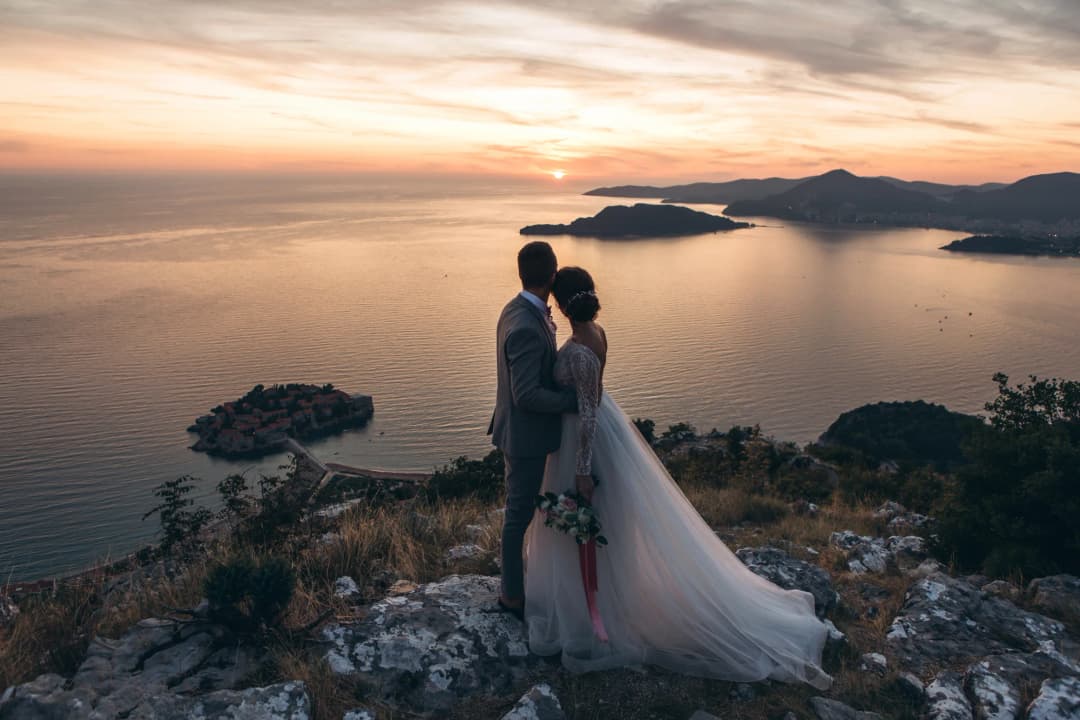 An elegant evening wedding reception in the courtyard of a stone palazzo in the Bay of Kotor, with string lights overhead, tables set with white linen, and the bay visible through an archway