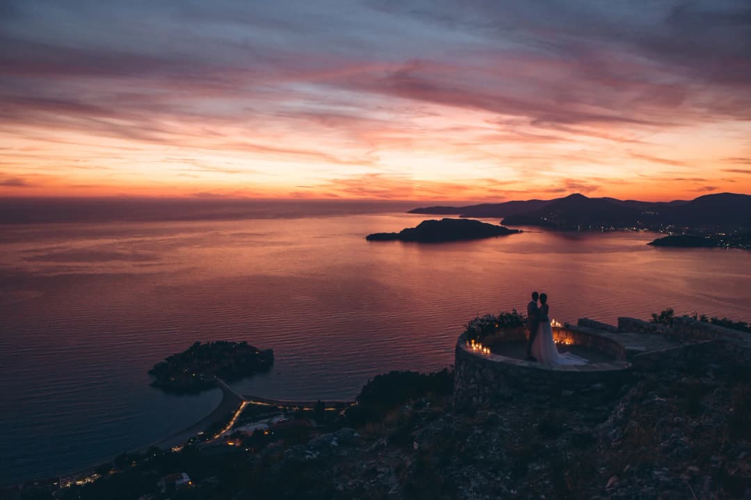 La fachada del hotel Iberostar Heritage Grand Perast al atardecer, con la terraza frente al agua iluminada preparada para una cena de boda y la Bahía de Kotor reflejando la última luz
