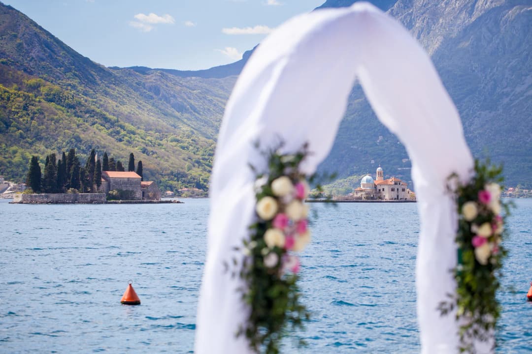 A wedding ceremony setup on a waterfront terrace in Perast, with white chairs facing the Bay of Kotor and Our Lady of the Rocks island visible in the background, golden hour light