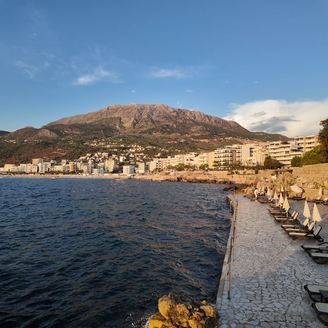 Plaža Veliki Pijesak i stambene zgrade Dobre Vode pod golom planinom, snimljene preko vode s hotelske terase sa ležaljkama u kasnom suncu