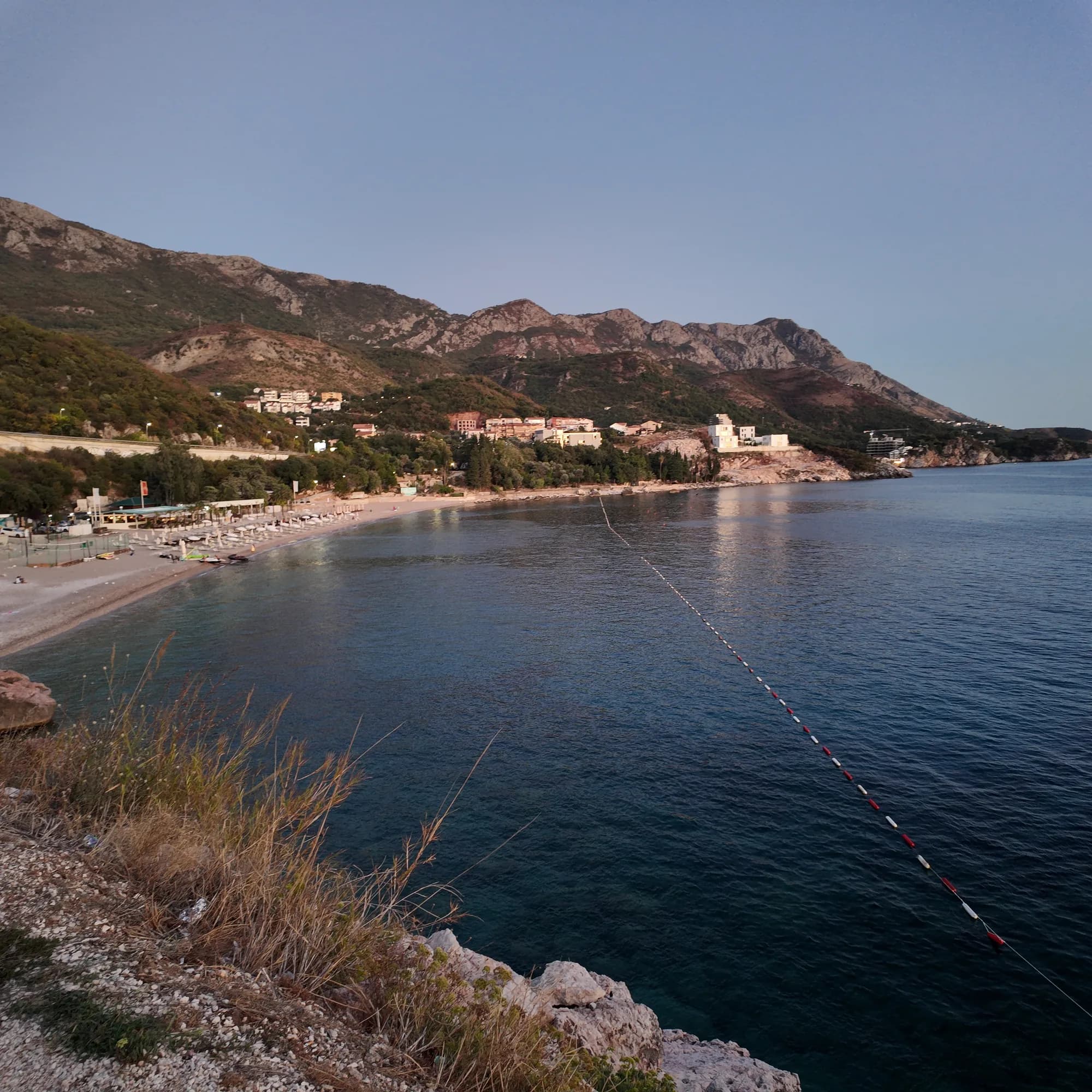 The full arc of Kamenovo beach from the coastal path on the Rafailovići side, buoyed bathing water and the hills of Pržno beyond