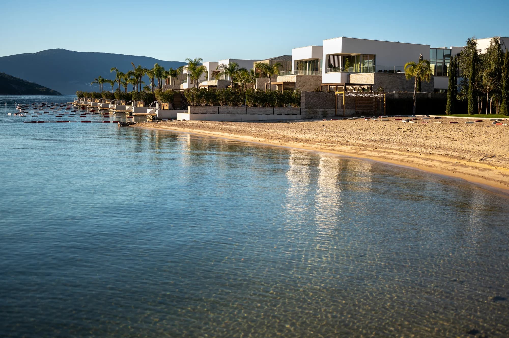 The sandy Kumbor beach at golden hour — clear shallows and palms along the waterfront