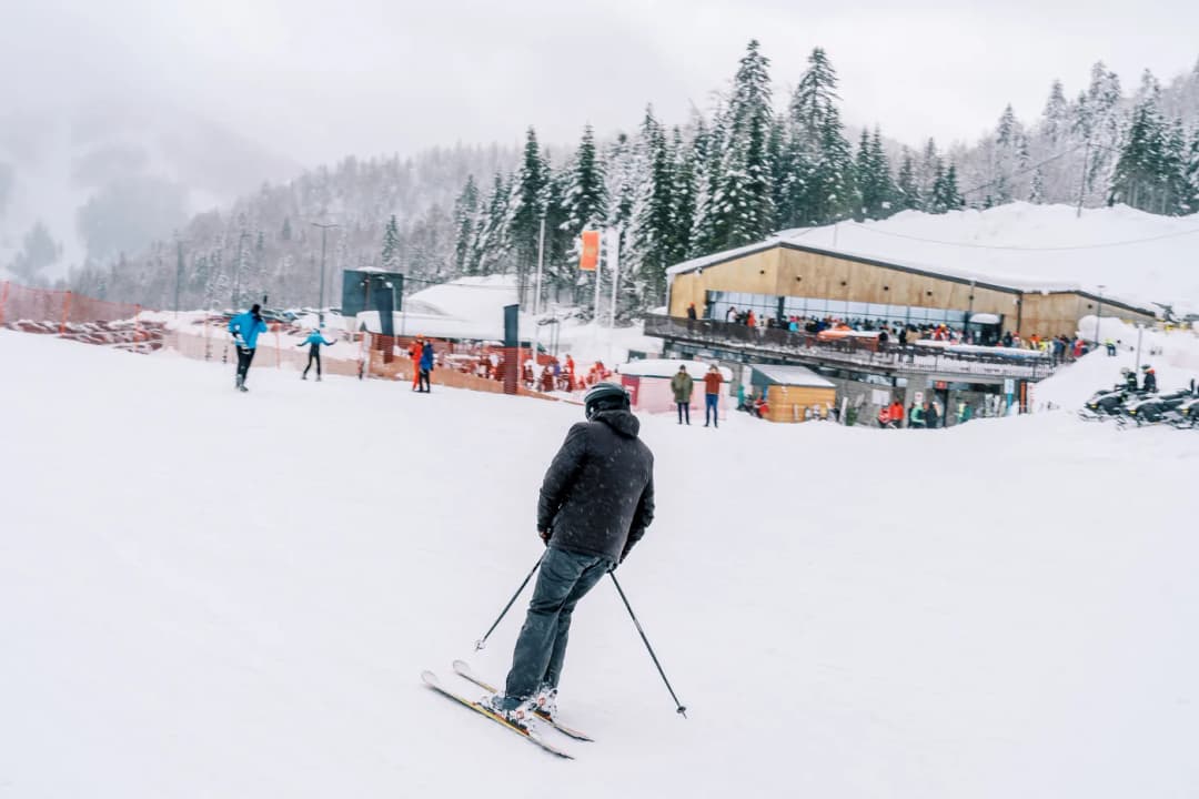 Skiers spread across a groomed piste in front of a timber resort lodge under falling snow.