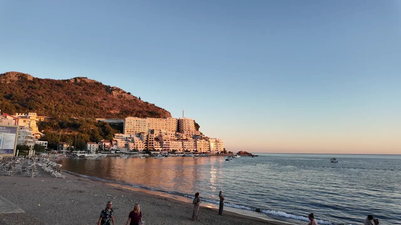 The Rafailovići end of the strand at sunset: the sand, boats at the mooring, the apartment blocks on the headland lit orange and the rock at the point