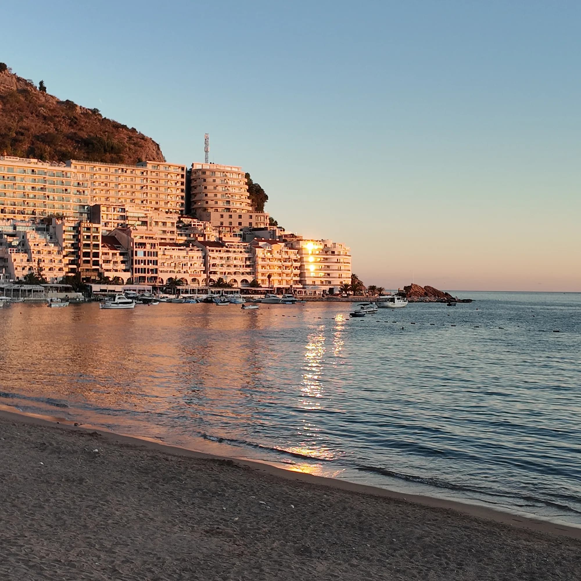 The Rafailovići end of the strand at sunset: the sand, boats at the mooring, the apartment blocks on the headland lit orange and the rock at the point