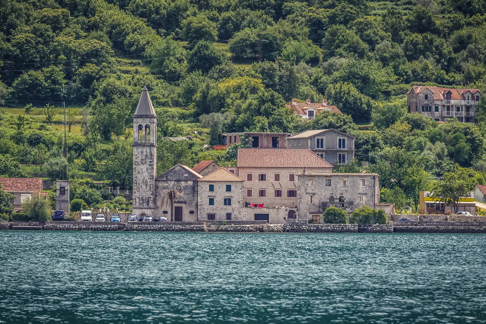 The Donji Stoliv waterfront from the water: the campanile and nave of the church of the Name of Mary, stone houses and the sea wall with cars parked on the shore road