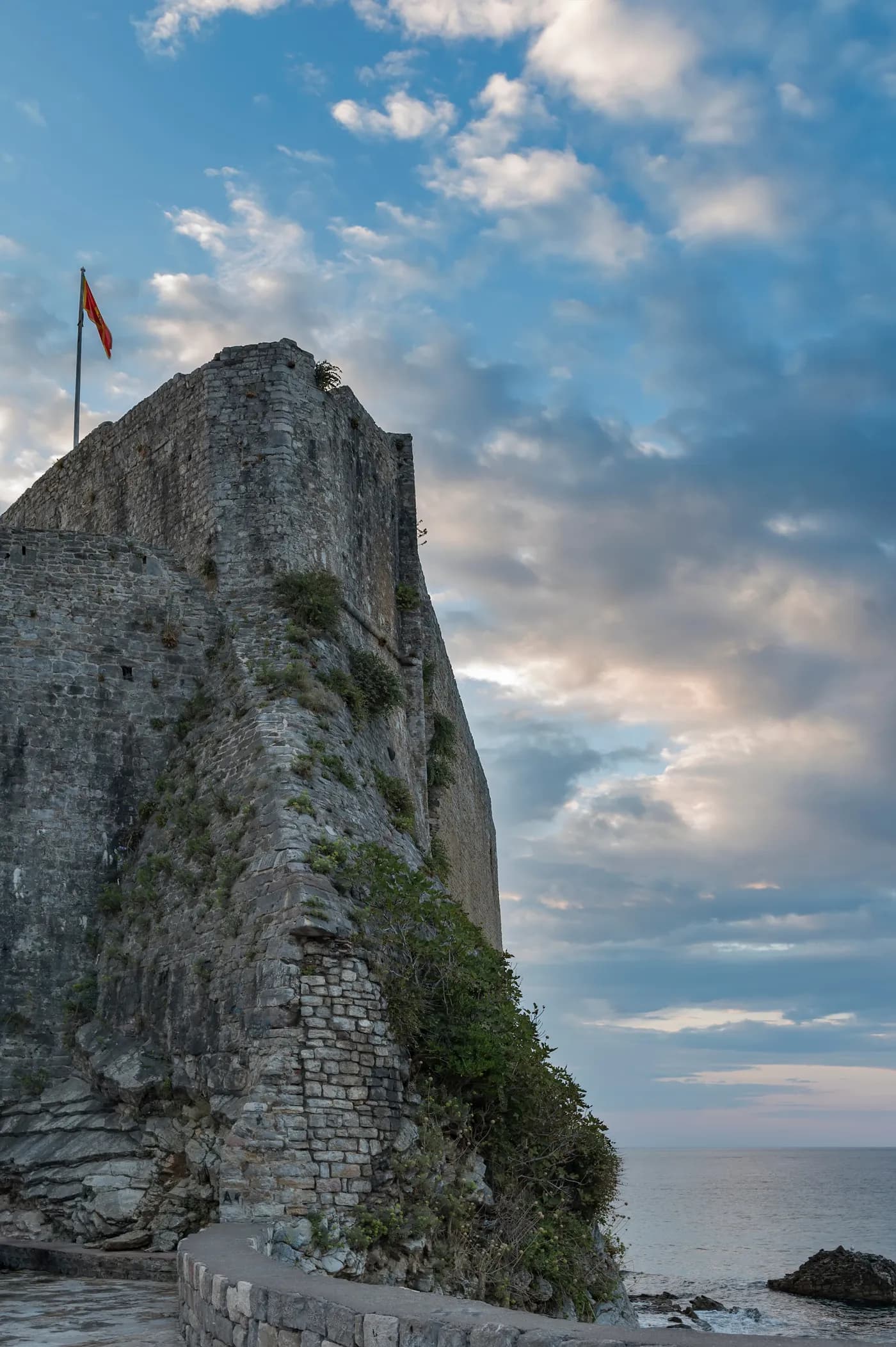 The Budva citadel from below at dusk: a massive battered stone wall rising from bare rock with a Montenegrin flag flying from the summit, vegetation growing from the joints, a stone buttress at its foot, and the calm open sea to the right.