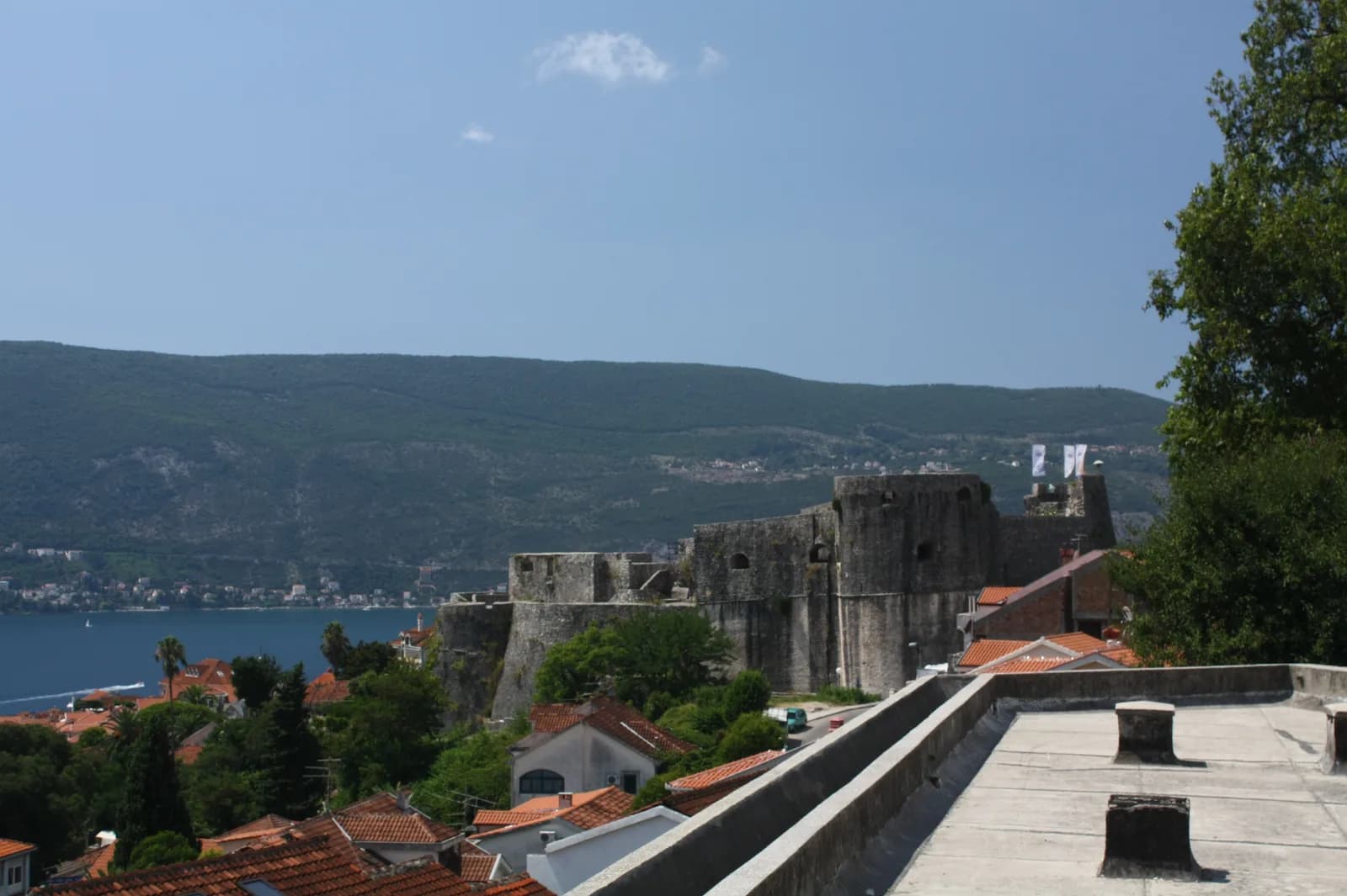 Kanli Kula seen from the south: a long grey stone fortress wall with a rounded bastion, standing above the red roofs of Herceg Novi with the Bay of Kotor and its far shore behind.