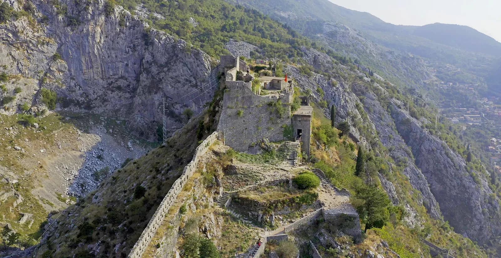 Aerial view of the fortress of San Giovanni above Kotor: stone buildings and a Montenegrin flag on the summit ridge, a crenellated curtain wall running down the spine of the rock, switchback steps climbing to it, a grey cliff face rising behind, and the valley far below at right.