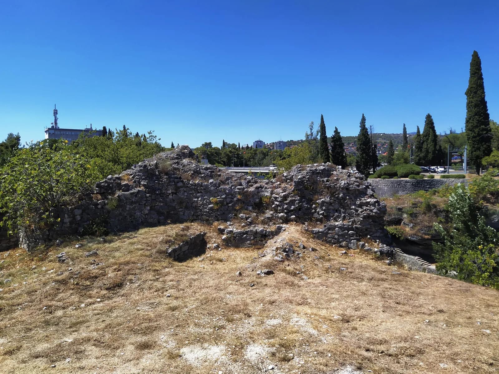 The ruined wall of the Ribnica fortress in Podgorica: a low, broken rubble-cored wall on dry grass, with a road bridge, trees, an office block and a telecommunications mast of the modern city visible behind it under a deep blue sky.