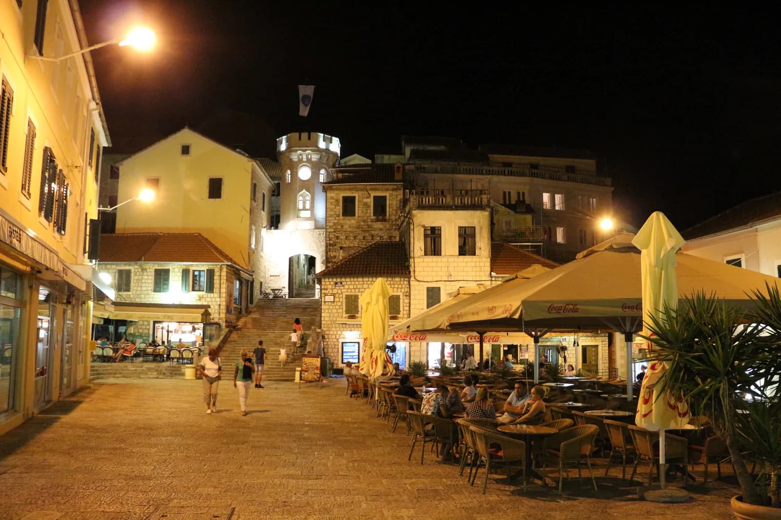 The Sahat kula lit at night at the top of a broad flight of stone steps, its crenellated top and clock face glowing above the roofs, with café tables and umbrellas filling the square in the foreground.