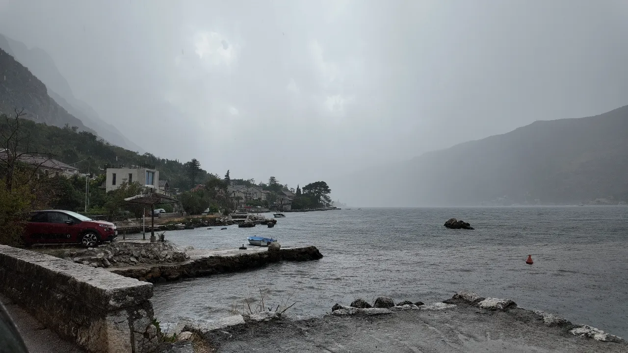 La lluvia barre la bahía de Kotor en Dobrota, con la carretera de la orilla mojada y un embarcadero delante, y la otra orilla perdida entre las nubes