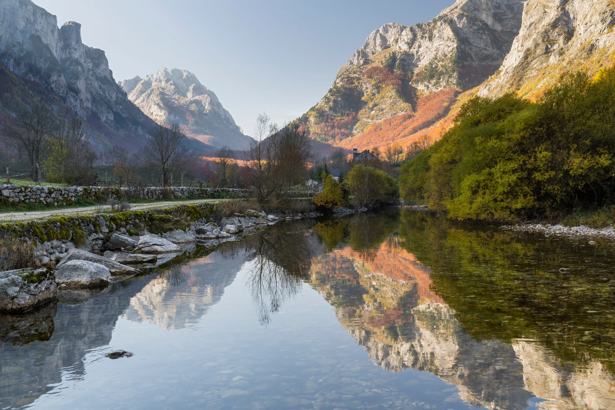 Un río de montaña cristalino que refleja picos calizos, con un muro de piedra seca y un camino de grava por la orilla
