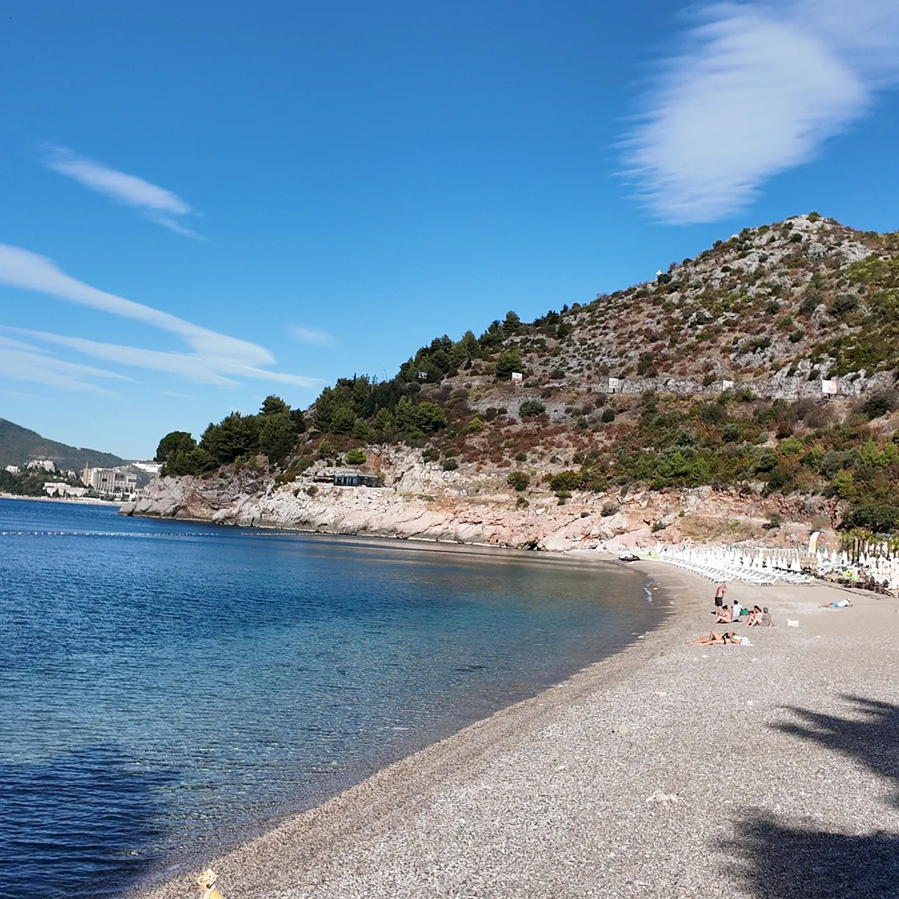 The full arc of Kamenovo beach in morning sun, umbrellas still closed, the buoy line across the bay and the hills of Pržno