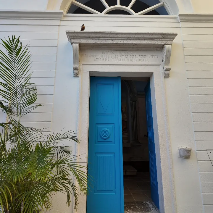 Die weiße Front und die blaue Doppeltür der Kapelle im Lazarett von Meljine, eine Inschrift über dem Türsturz und eine Palme daneben