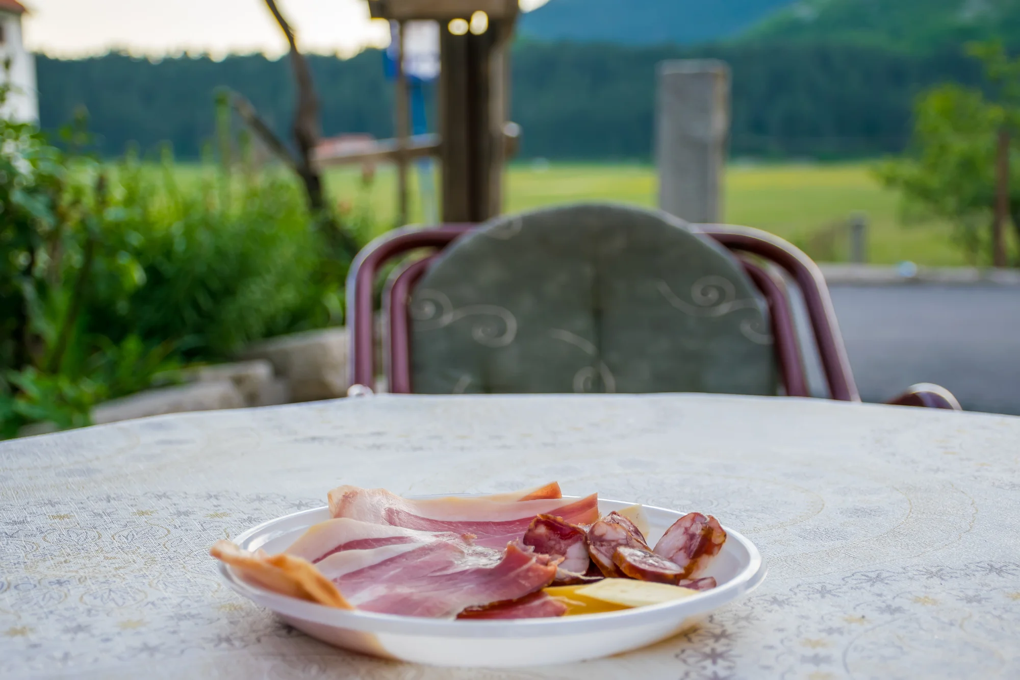 Un plato de pršut en lonchas finas, embutido curado y queso en una mesa al aire libre, con un campo verde y la linde del bosque al fondo