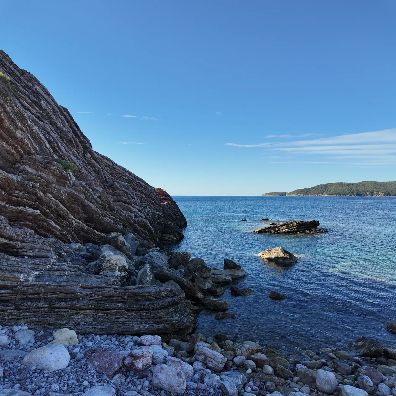 Layered red-brown rock of Cape Đevištenje standing in the sea, flat blue water and the island of Sveti Nikola in the distance