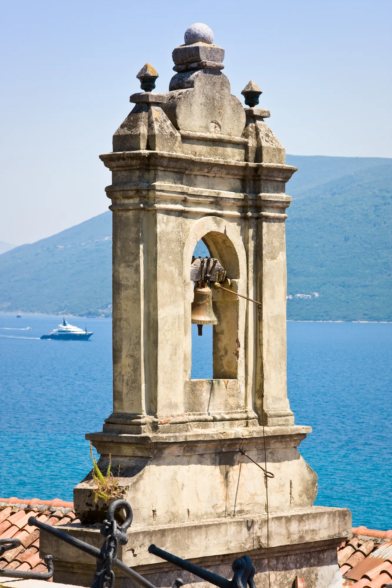 A weathered stone bell-cote with a single bell, the bay and a passing yacht far below