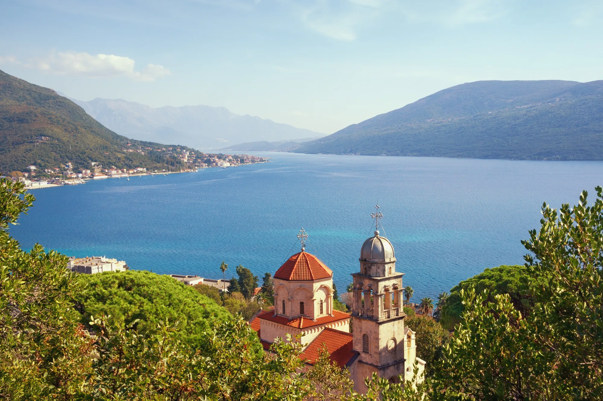 The monastery church half-hidden in greenery on the shoulder above the bay, the strait running out toward the sea between mountain shores