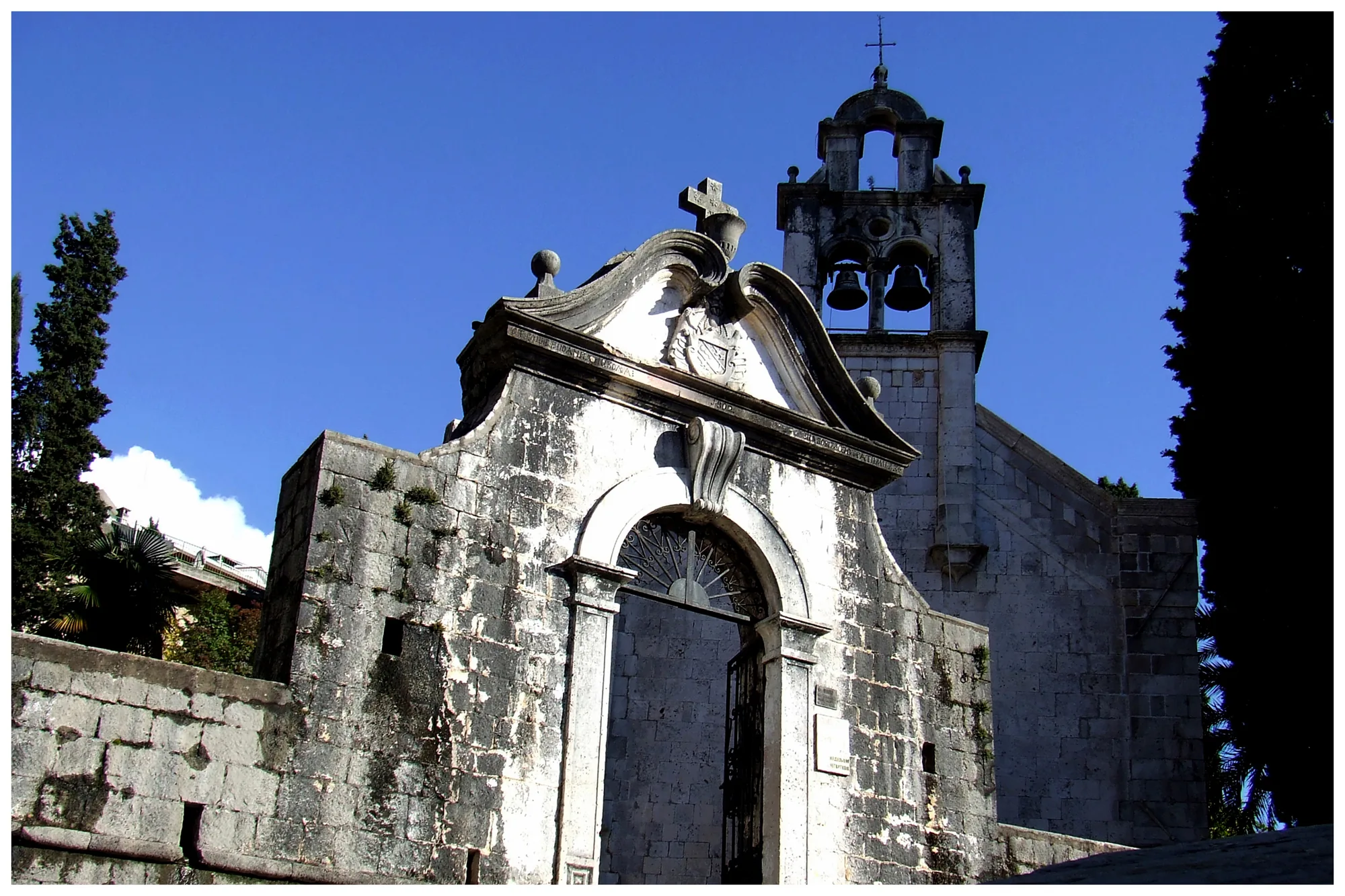 A baroque stone gateway with a scrolled pediment and a cross, a triple bell-cote rising behind it against a clear sky