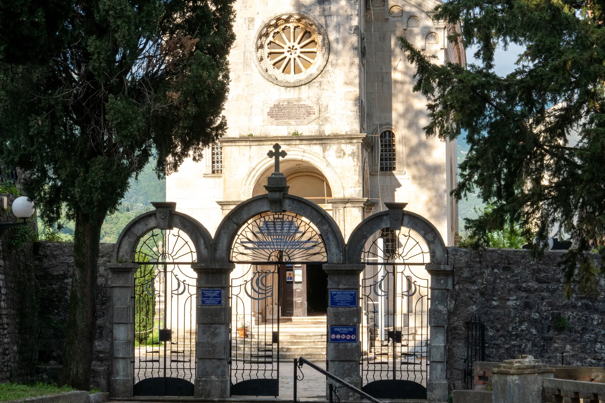 A church front with a rose window and a bell tower behind it, seen through the ironwork gates of its walled courtyard, cypresses on either side