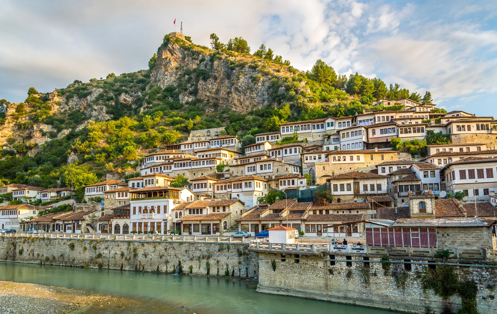 The white Ottoman houses of Berat, Albania, stacked on a hillside below a rocky ridge, seen from across the Osum river